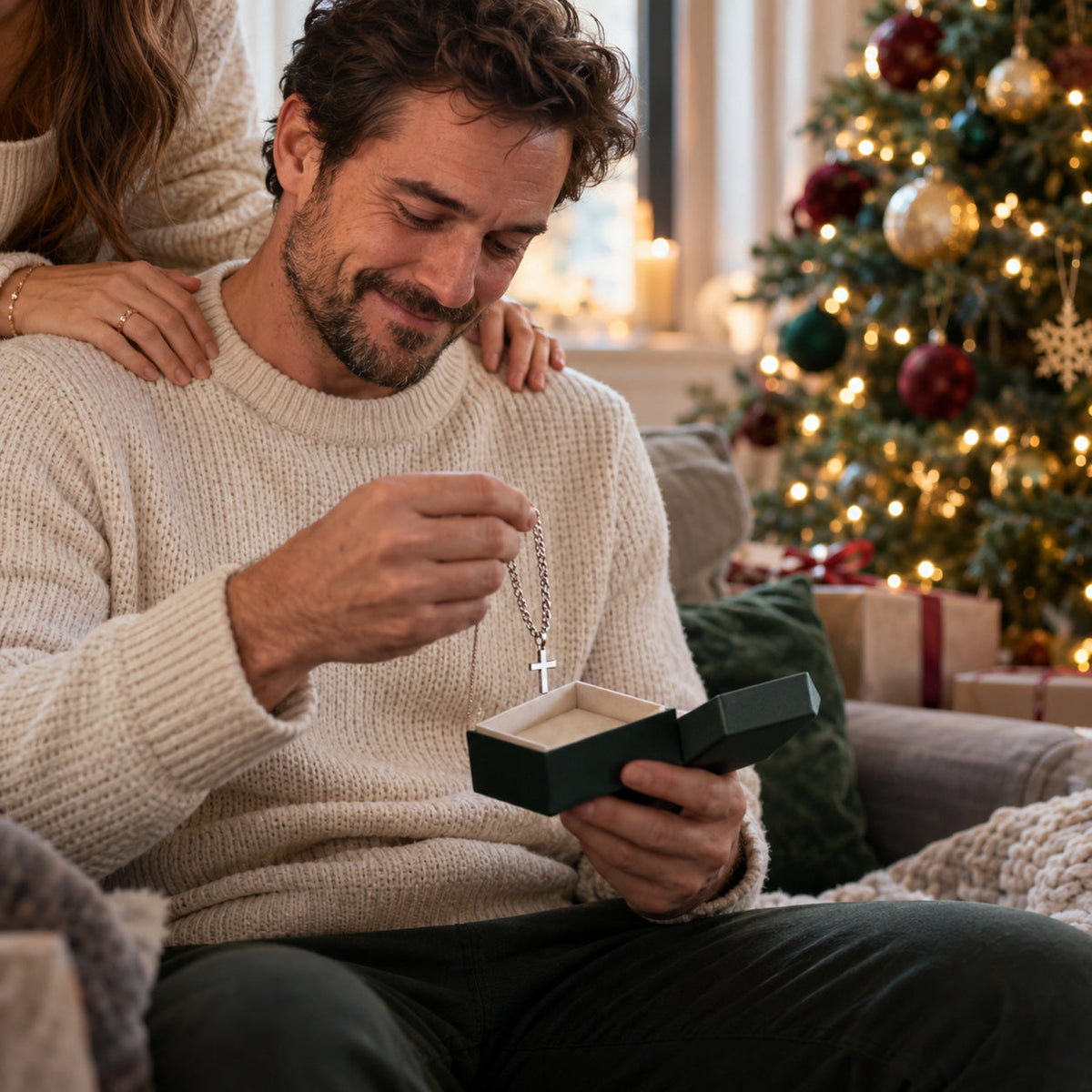 Man smiling as he opens a Christmas gift box with a silver cross necklace, wife's hands on his shoulders – Christian Christmas gifts for husband