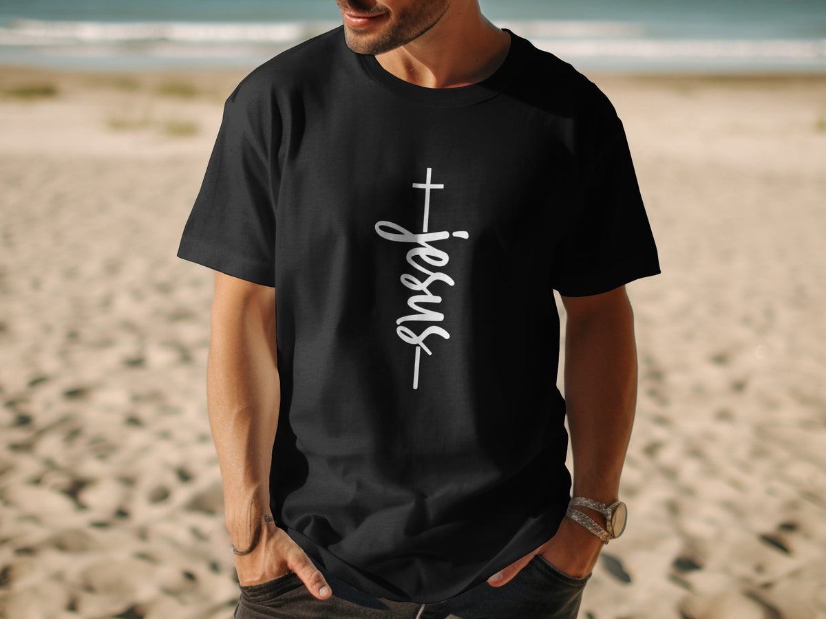 Black t-shirt with white "Jesus" text and cross on chest, model on beach.